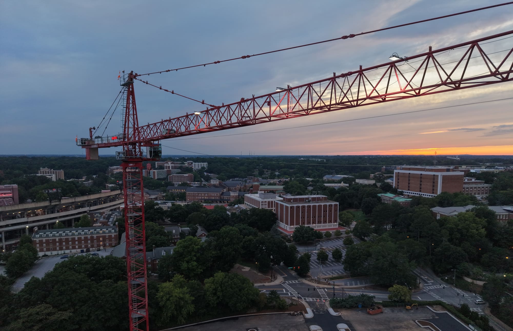 A tower crane over a campus construction site at dusk, with the sun setting behind the treeline