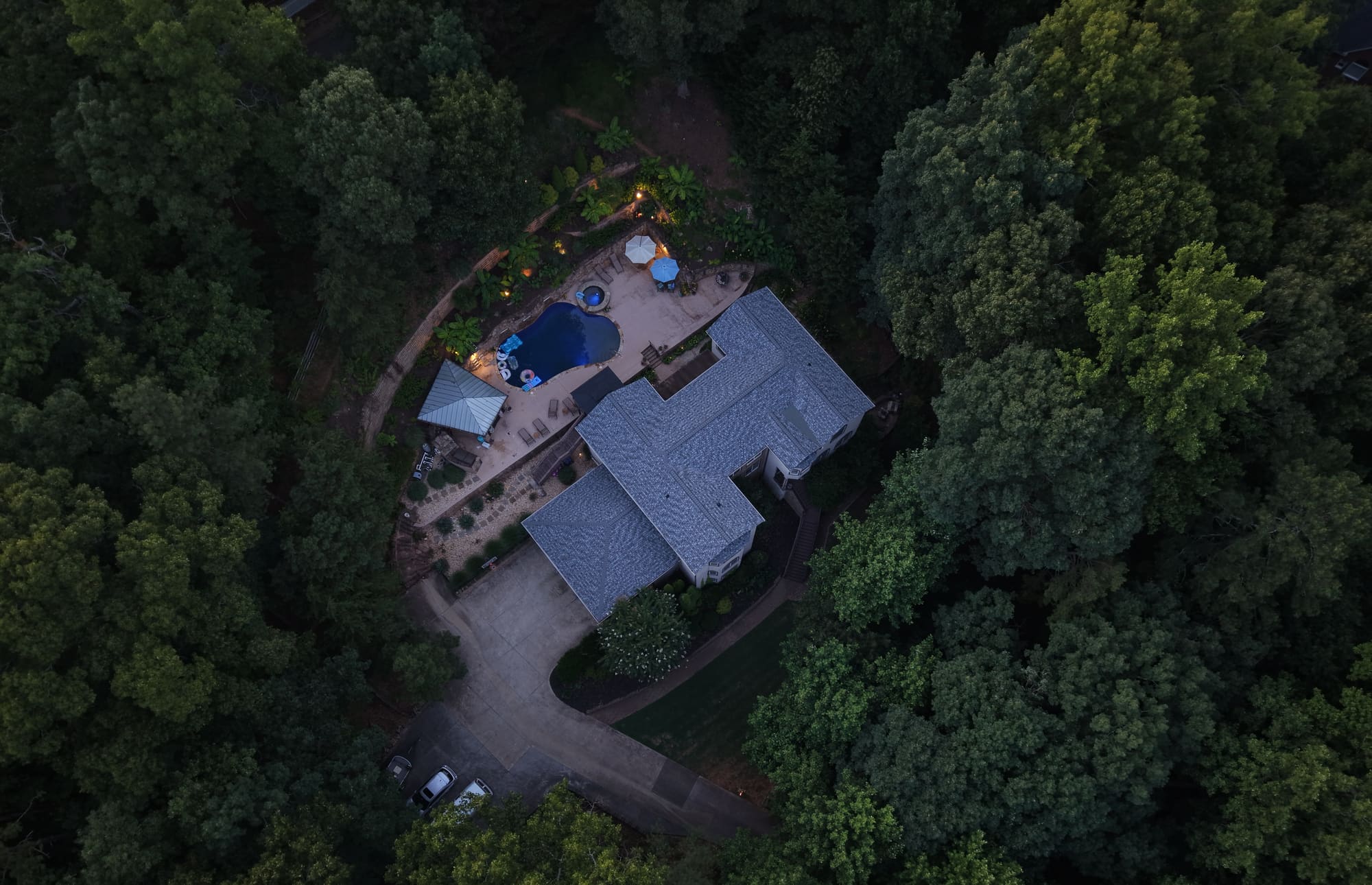 Overhead twilight view of a house in the trees, with a lit pool and patio beside it