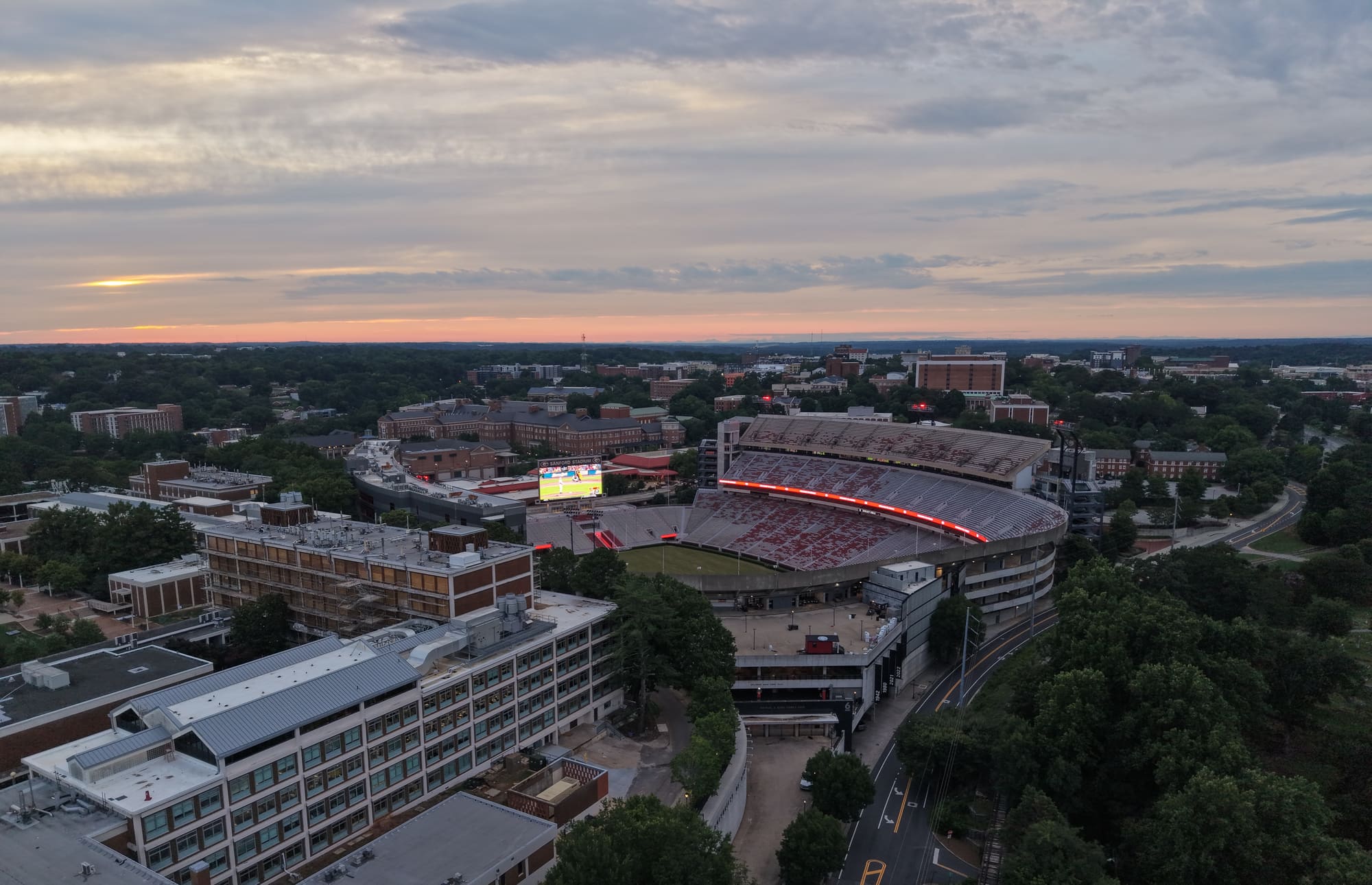 A college football stadium at dusk, lit scoreboard on, with the campus around it