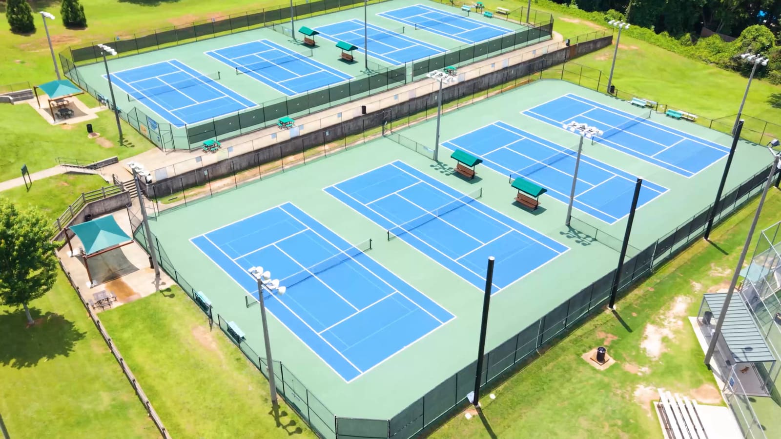 Aerial view of two banks of blue tennis courts at Terrell Mill Park, with floodlight poles and mown grass around them